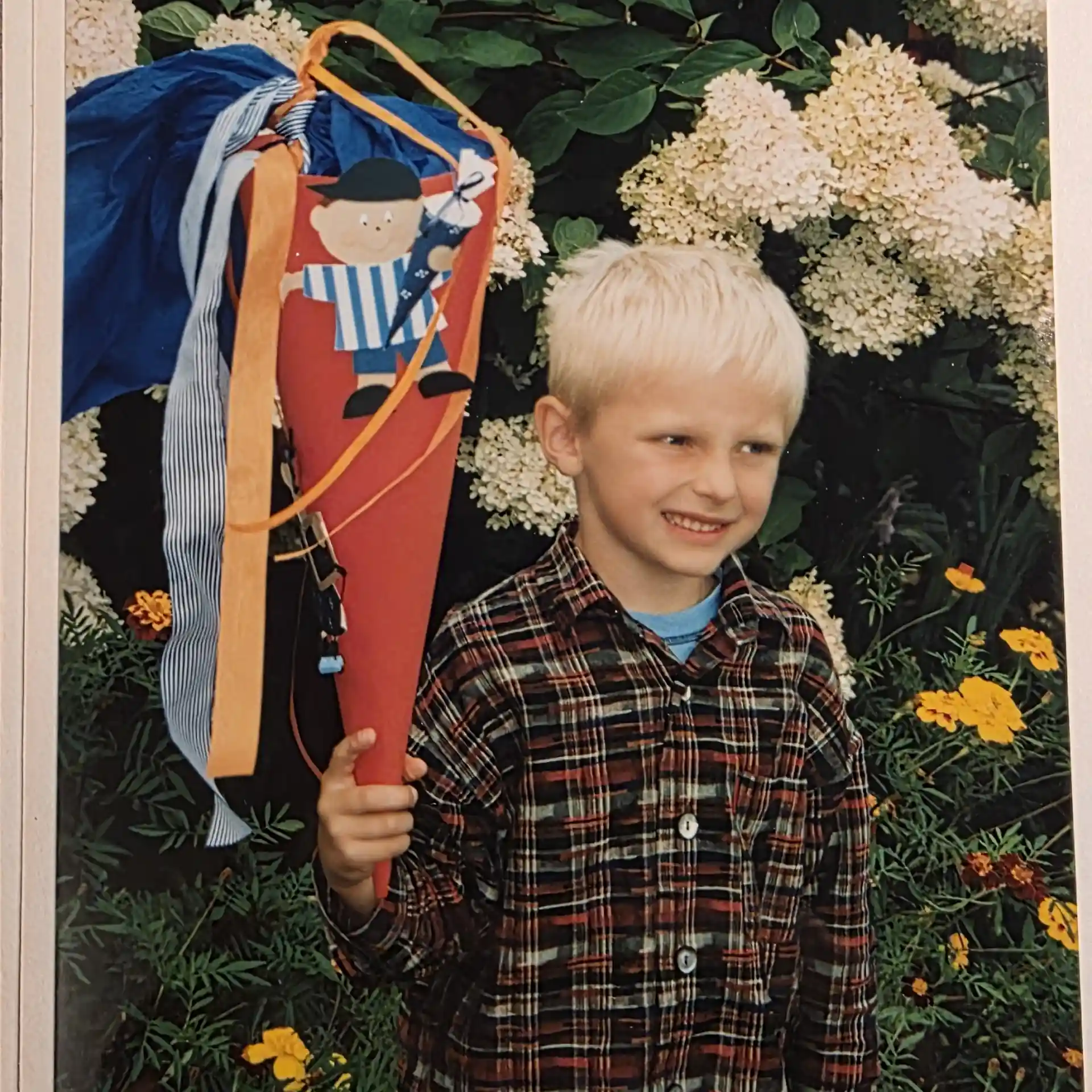 Julian mit Schultüte Julian als kleines Kind mit einer Schultüte in der Hand vor Blumen stehend.